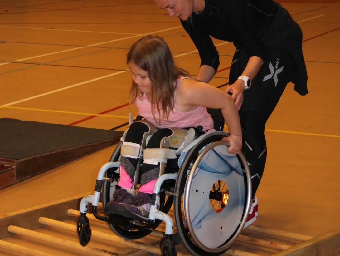 En jente øver på å kjøre rullestolen over en rist. Instruktør går bak for veiledning og sikkerhet