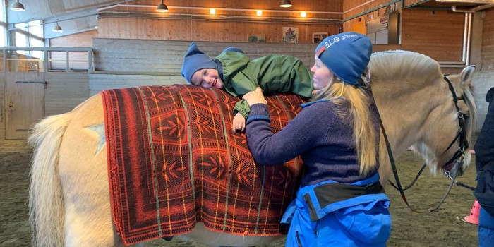En smilende gutt ligger på magen på hesteryggen, mens rideinstruktøren holder han fast